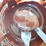 A metal fork holds a crunchy peanut butter egg coated with chocolate over a bowl of melted chocolate, with smears on the sides.