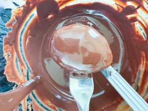 A metal fork holds a crunchy peanut butter egg coated with chocolate over a bowl of melted chocolate, with smears on the sides.
