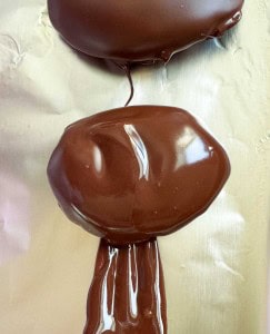 Two oval-shaped crunchy peanut butter eggs coated in melted chocolate rest on a sheet of foil, with excess chocolate dripping downward.
