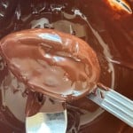 A close-up of a metal spoon coated in melted chocolate, with crunchy peanut butter eggs and more melted chocolate visible in the bowl underneath.