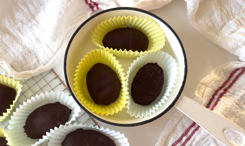 A white pan holds three crunchy peanut butter eggs in yellow paper cups, surrounded by more chocolate-covered treats on a cooling rack and white towels.