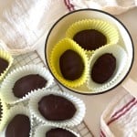 Crunchy peanut butter eggs coated in chocolate are arranged in paper liners on a cooling rack and inside a white pan, with white towels surrounding them.