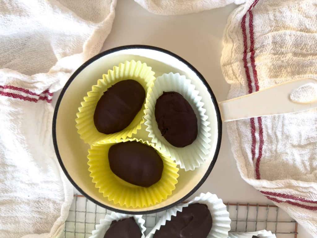 A small white pan containing chocolate-coated, crunchy peanut butter eggs in paper liners, placed on a white surface with red-striped towels nearby.