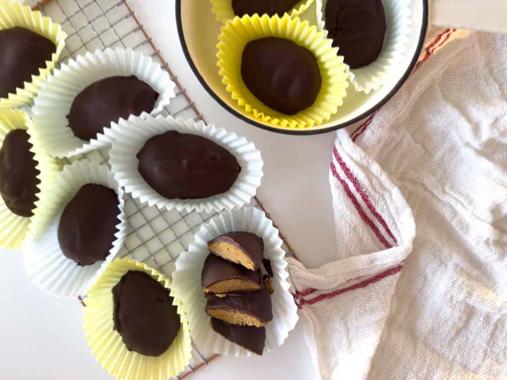 Chocolate-covered treats, including some crunchy peanut butter eggs&mdash;both whole and halved to reveal their filling&mdash;are arranged in paper liners on a cooling rack and in a bowl beside a white towel with red stripes.