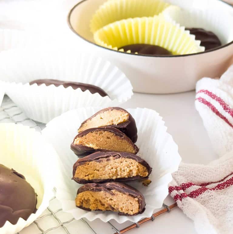 Three crunchy peanut butter eggs coated in chocolate are cut in half and arranged in a white cupcake liner, with more whole eggs in paper liners and a dish visible in the background.