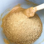Crushed graham crackers being mixed into a creamy brown batter with crunchy peanut butter eggs using a spatula inside a light-colored mixing bowl.