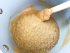 Crushed graham crackers being mixed into a creamy brown batter with crunchy peanut butter eggs using a spatula inside a light-colored mixing bowl.