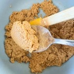 Spoon holding a scoop of crunchy peanut butter cookie dough above a mixing bowl, with a spatula resting on the side&mdash;perfect for making delicious peanut butter eggs.