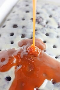 Caramel sauce being poured over a Heath Bar Poke Cake with holes and a white glaze, creating an irresistible dessert.