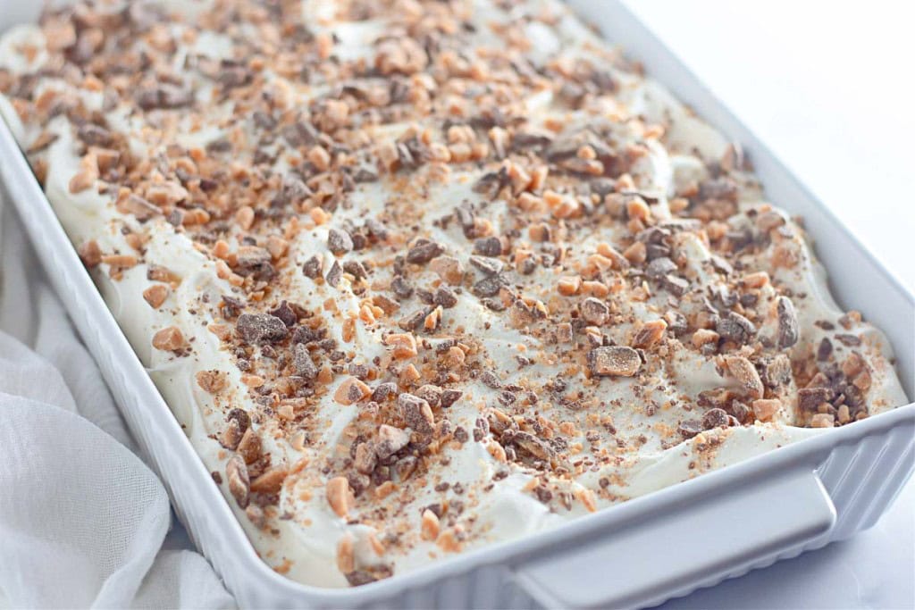 A rectangular dish filled with Heath Bar Poke Cake, topped with whipped cream and crushed toffee pieces.