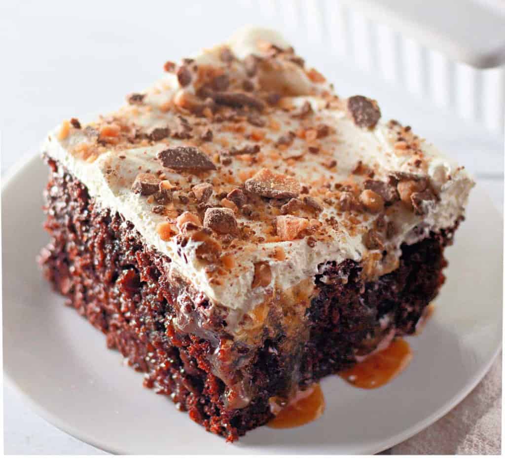 A slice of Heath Bar Poke Cake topped with whipped cream, caramel sauce, and crushed candy pieces sits on a white plate.