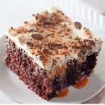 A slice of Heath Bar Poke Cake topped with whipped cream, caramel sauce, and crushed candy pieces sits on a white plate.