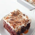 A slice of Heath Bar Poke Cake topped with whipped cream and crushed candy pieces, served on a white plate with caramel sauce visible.