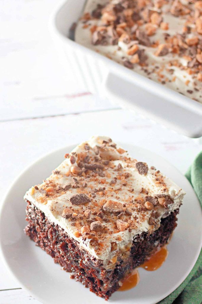 A slice of Heath Bar Poke Cake topped with whipped cream and crushed toffee, served on a white plate with caramel sauce, sits next to a baking dish filled with more cake.