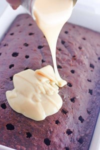 Sweetened condensed milk is being poured from a can over a Heath Bar Poke Cake, filling the holes in its rich chocolate surface.
