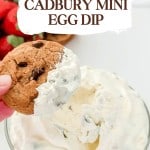 A hand dips a chocolate chip cookie into creamy Cadbury Mini Egg Dip, with strawberries in the background for a festive touch.