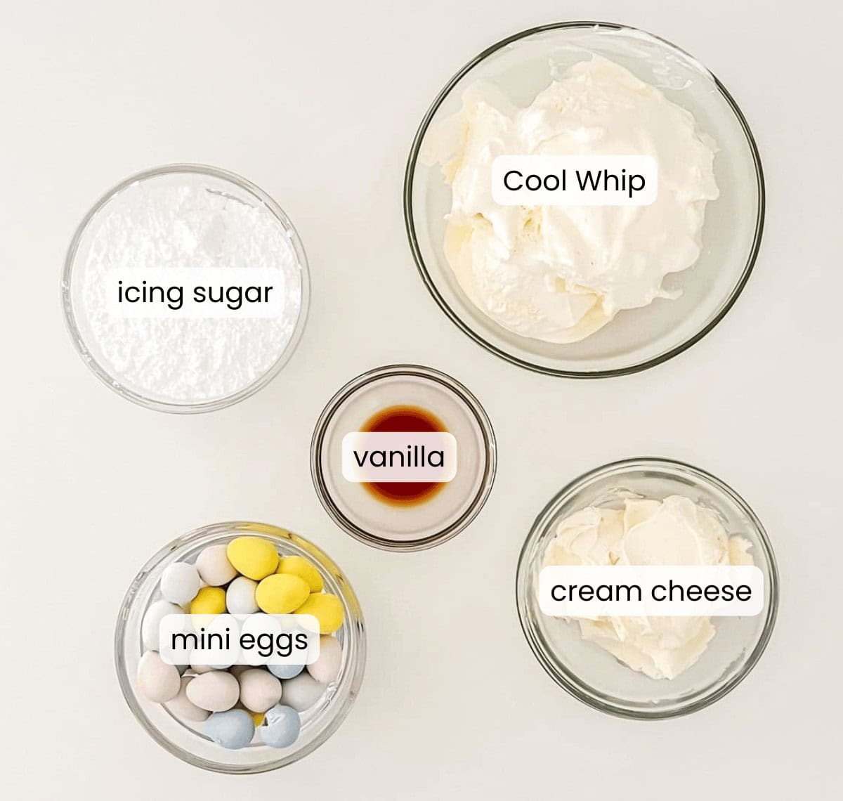 Five labeled bowls containing icing sugar, Cool Whip, cream cheese, mini eggs, and vanilla are arranged on a white surface&mdash;perfect ingredients for making a festive Cadbury Mini Egg Dip.