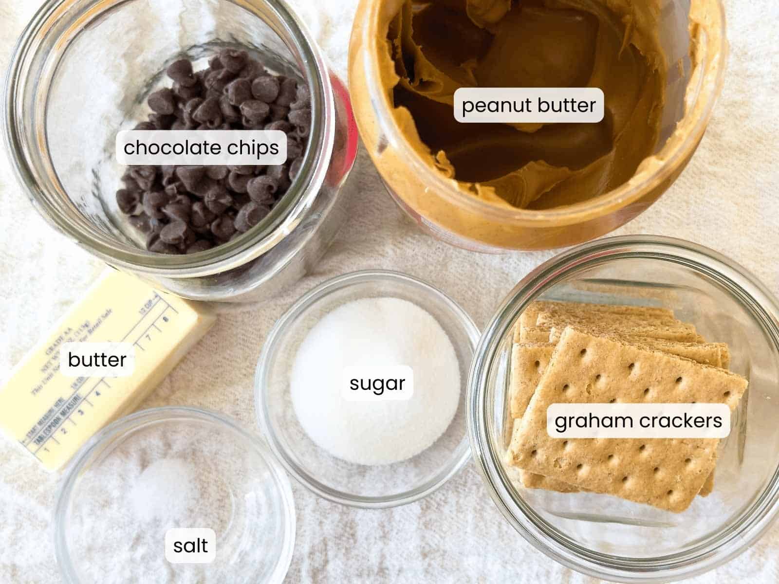 Top-down view of ingredients in separate containers: chocolate chips, crunchy peanut butter eggs, butter, sugar, salt, and graham crackers, each labeled with text.