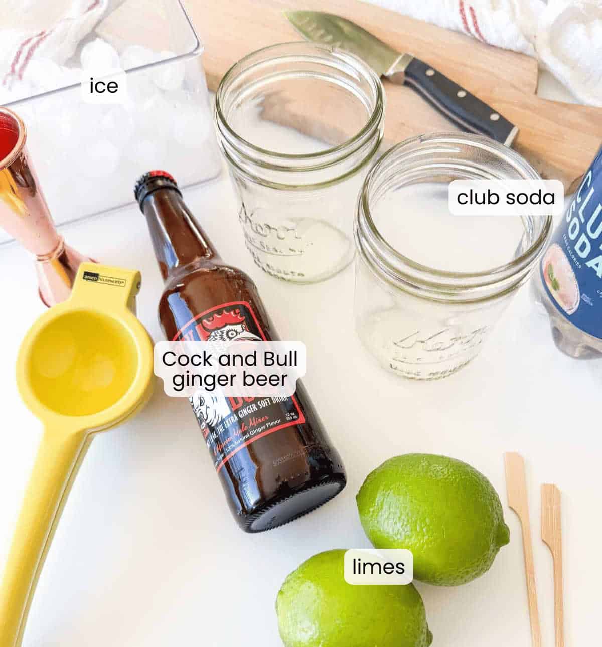 Top-down view of Mocktail Moscow mule ingredients, including a bottle of Cock and Bull ginger beer, club soda can, two limes, yellow citrus press, glass jars, ice, and cocktail picks arranged on a counter.
