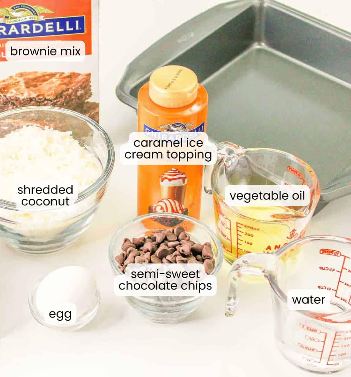 Samoa brownies ingredients&mdash;brownie mix, shredded coconut, egg, semi-sweet chocolate chips, caramel ice cream topping, vegetable oil, and water&mdash;are arranged in a baking pan on a white surface.