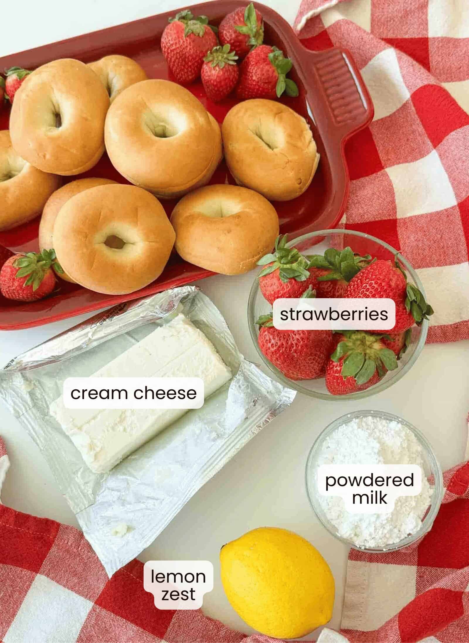 A red dish with plain bagels and strawberries, next to strawberry cream cheese, a bowl of strawberries, powdered milk, and a lemon on a red-and-white checkered cloth.