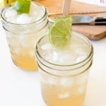 Two mason jars filled with iced Mocktail Moscow Mules, garnished with lime slices on wooden skewers, sit on a white surface with a cutting board and knife in the background.