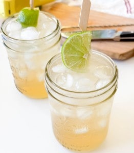 Two mason jars filled with iced Mocktail Moscow Mules, garnished with lime slices on wooden skewers, sit on a white surface with a cutting board and knife in the background.