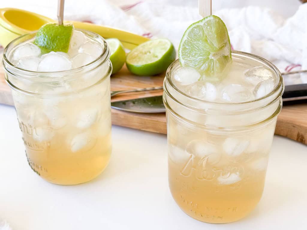 Two mason jar glasses filled with ice and light-colored Mocktail Moscow mule, garnished with lime wedges, sit on a white surface beside a cutting board with sliced limes and a knife.