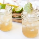 Two mason jar glasses filled with ice and light-colored Mocktail Moscow mule, garnished with lime wedges, sit on a white surface beside a cutting board with sliced limes and a knife.