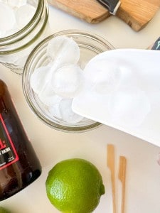 A white scoop adds round ice cubes to a glass jar for a refreshing Mocktail Moscow mule. Nearby are a lime, a brown bottle, wooden stir sticks, and a cutting board with a knife.