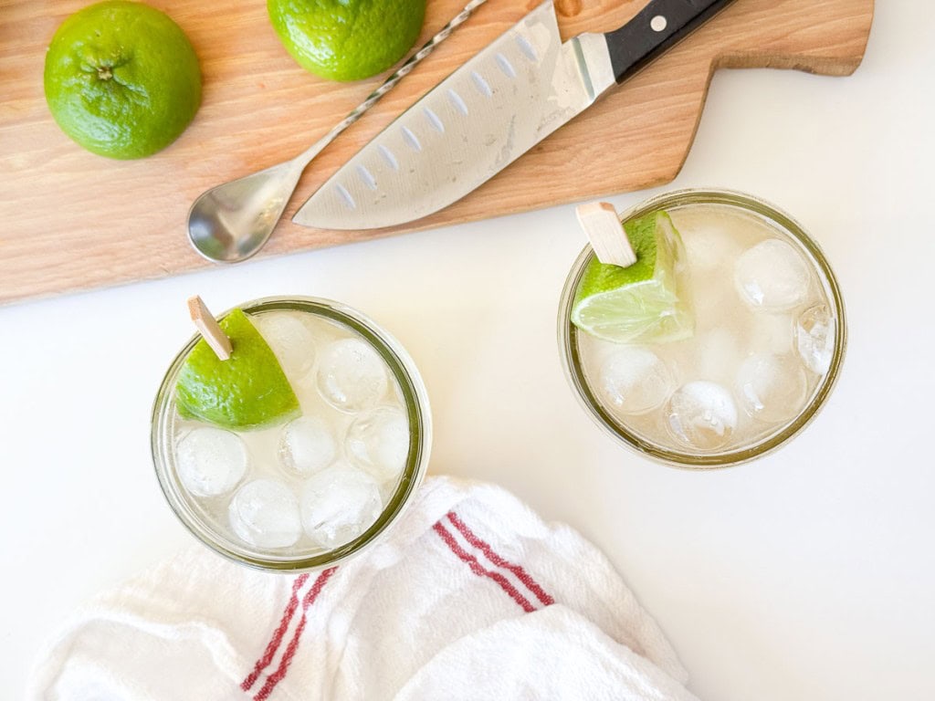 Two glasses with ice and lime wedges sit on a white surface beside a cutting board with limes, a knife, and a metal spoon; perfect for preparing a refreshing Mocktail Moscow mule. A white towel with a red stripe is nearby.