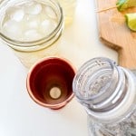 A clear glass of ice water, a copper cup ready for a Mocktail Moscow mule with a tablet inside, a plastic water bottle, and a wooden board with pieces of lime on a white surface.