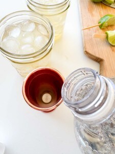 A clear glass of ice water, a copper cup ready for a Mocktail Moscow mule with a tablet inside, a plastic water bottle, and a wooden board with pieces of lime on a white surface.