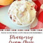 A bowl of strawberry cream cheese sits on a white plate next to a red and white checkered cloth and two bread rolls.