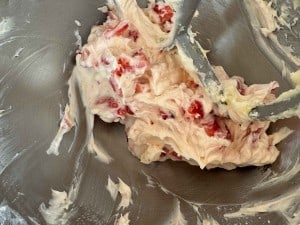 A close-up of a mixing bowl with partially mixed cream and chopped strawberries, showing a stand mixer attachment coated in the mixture.