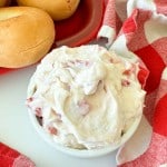 A small white bowl of creamy spread with bits of red pepper sits next to plain bagels and a red and white checkered cloth.