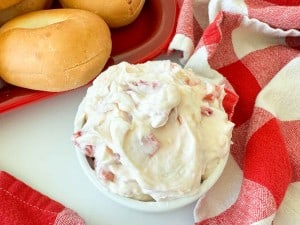 A small white bowl of creamy spread with bits of red pepper sits next to plain bagels and a red and white checkered cloth.