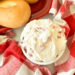 A bowl of cream cheese with pieces of strawberry next to plain bagels on a red tray and a red-and-white checkered cloth.