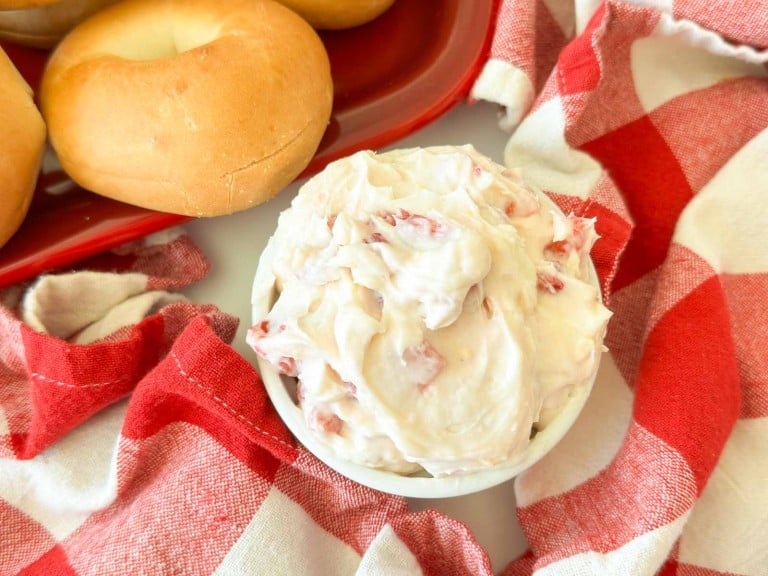 A bowl of cream cheese with pieces of strawberry next to plain bagels on a red tray and a red-and-white checkered cloth.
