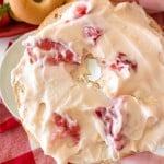 A hand holds a bagel half topped with strawberry cream cheese spread; whole bagels and a red checkered cloth are in the background.