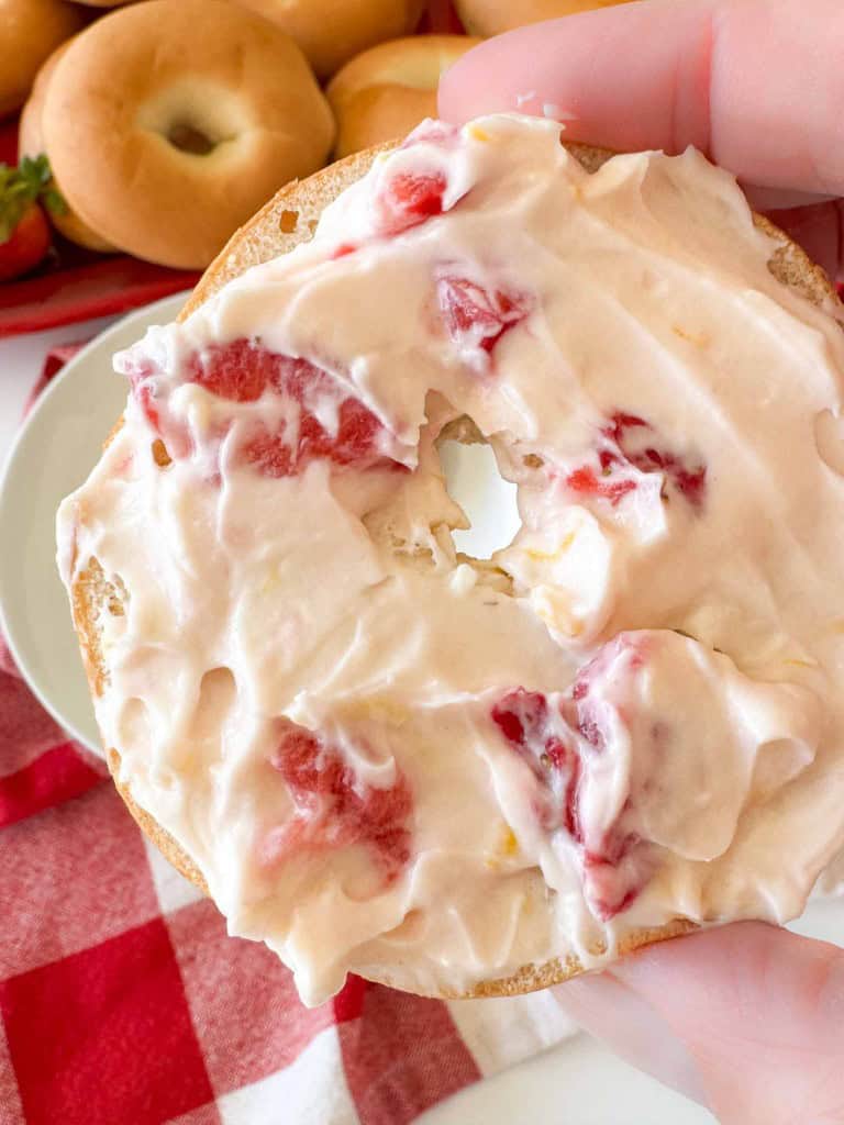 A hand holds a bagel half topped with strawberry cream cheese spread; whole bagels and a red checkered cloth are in the background.