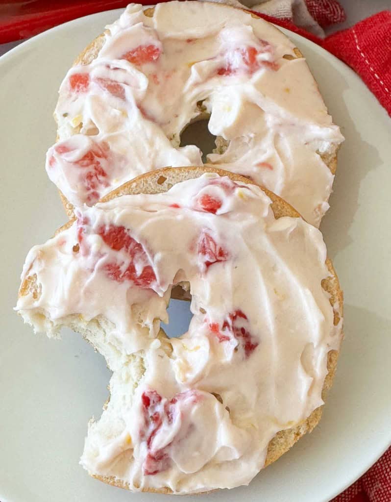 Two bagel halves topped with strawberry cream cheese are on a white plate, with a bite taken out of one half.