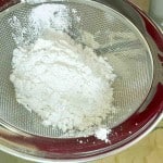 A metal sieve with white flour inside positioned over a mixing bowl containing more flour.