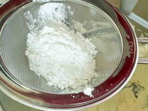 A metal sieve with white flour inside positioned over a mixing bowl containing more flour.