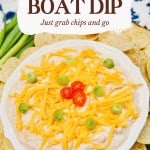 A bowl of creamy dip topped with shredded cheddar cheese, sliced green onions, and cherry tomatoes, surrounded by round tortilla chips.