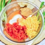 A glass bowl with shredded cheese, diced tomatoes, sour cream, seasoning, and sauce on a wooden board, surrounded by green onions and tortilla chips.