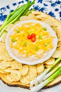 A bowl of dip topped with shredded cheese, sliced green onions, and red chili, surrounded by round tortilla chips and fresh green onions on a wooden platter.