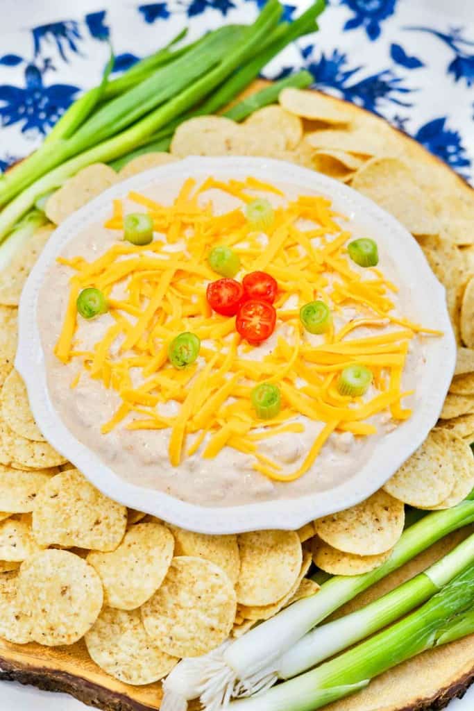 A bowl of creamy dip topped with shredded cheddar cheese, sliced green onions, and cherry tomatoes, surrounded by tortilla chips and fresh green onions.
