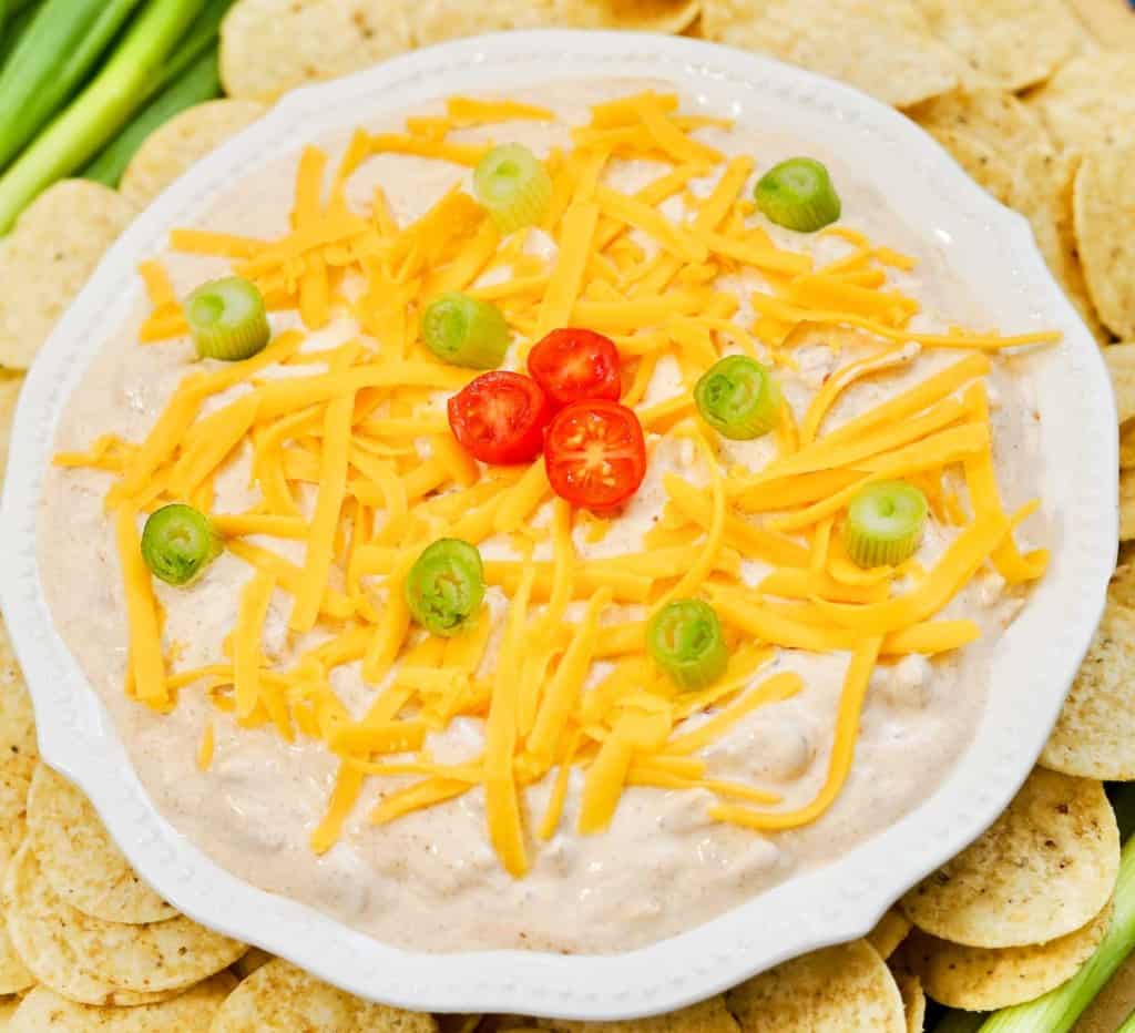A plate of creamy dip topped with shredded cheddar cheese, sliced green onions, and cherry tomato halves, surrounded by tortilla chips.