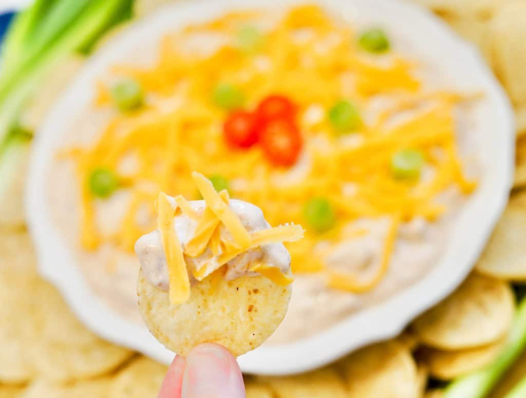 A hand holds a chip topped with dip and shredded cheese, with a plate of dip garnished with cheese, tomatoes, and green onions in the background.